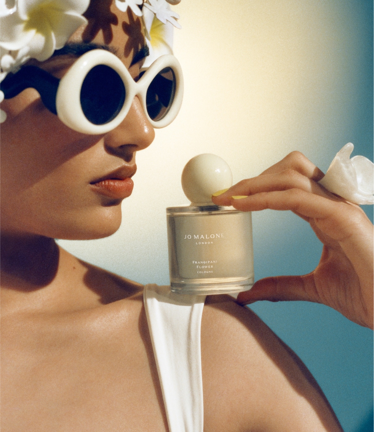 A person wearing white swimwear and sunglasses, holds a Jo Malone London cologne on their shoulder against a blue background. 