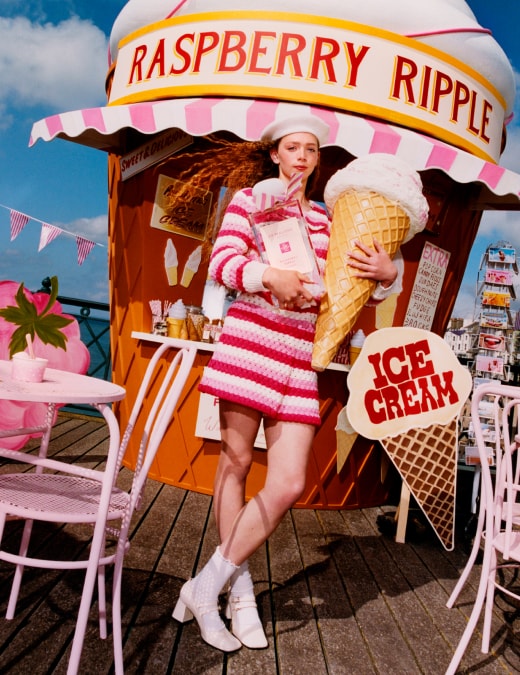 A person in pink holds an oversized ice cream and Jo Malone London cologne on a pier, with pink seating, and a giant cone hut.