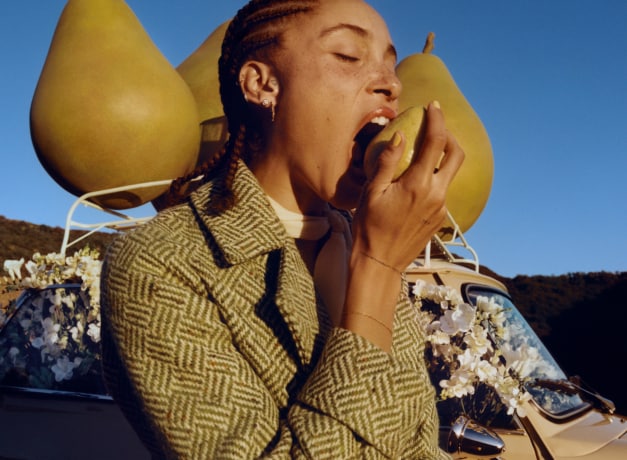 Adwoa Aboah with braided hair, biting into a pear with backdrop of three large pear props placed on top of a car roof rack.